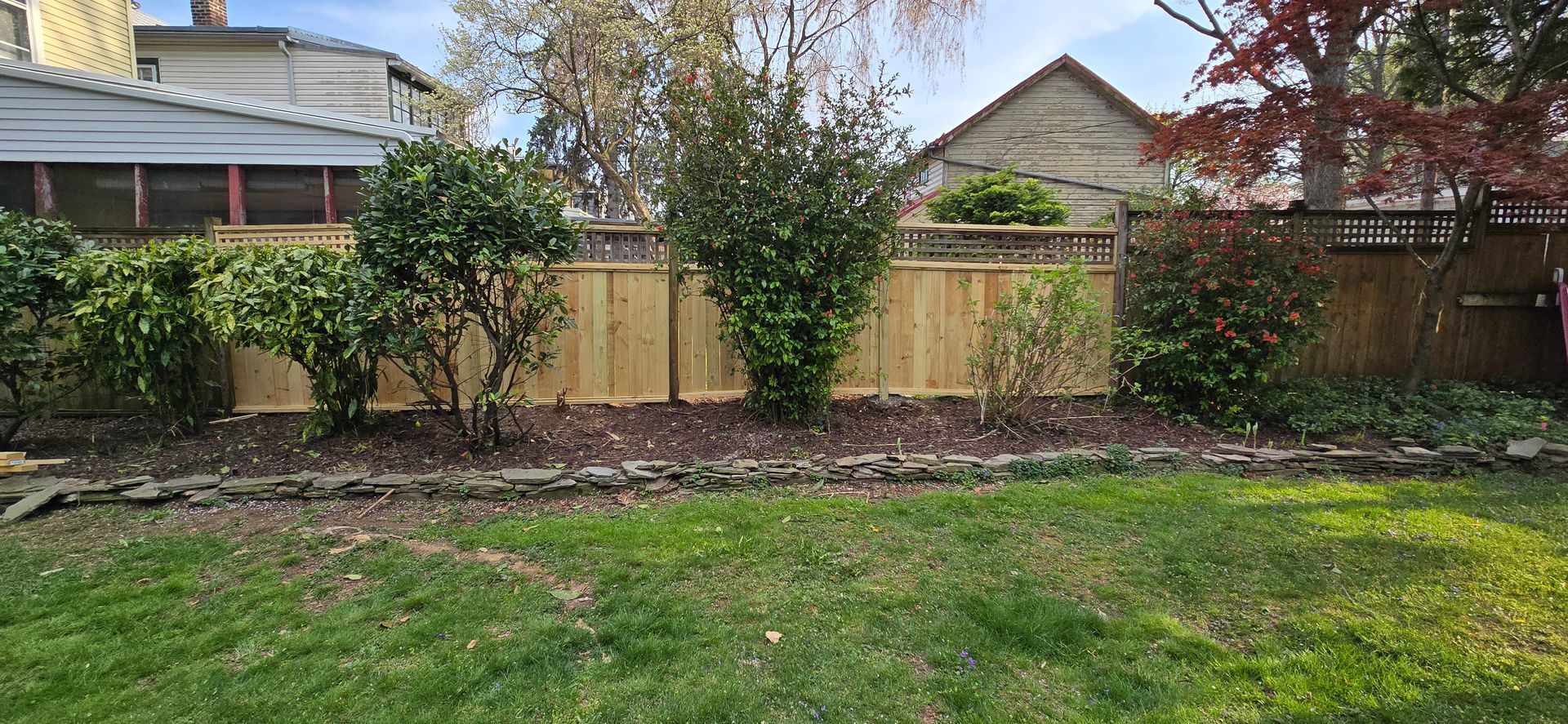 Backyard with fence, landscaping, American flag on pole, blue sky.