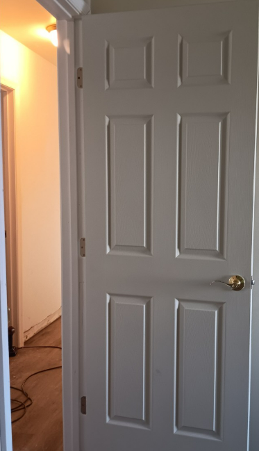 White six-panel interior door, slightly ajar, doorway to a lit hallway.