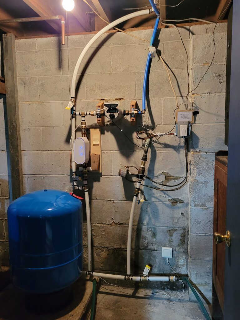 Plumbing system mounted on a gray concrete block wall. Blue water tank, pipes, and valves are visible.